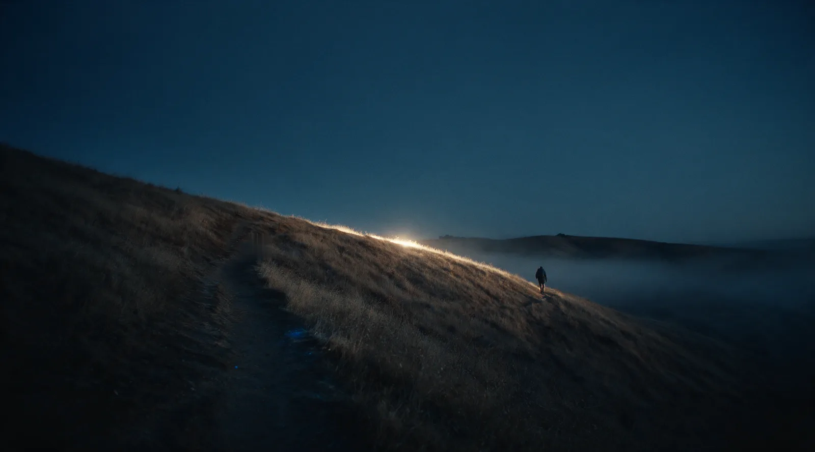 A dirt trail climbing a grassy ridge before sunrise, a distant hiker ahead, the first light breaking on the ridge